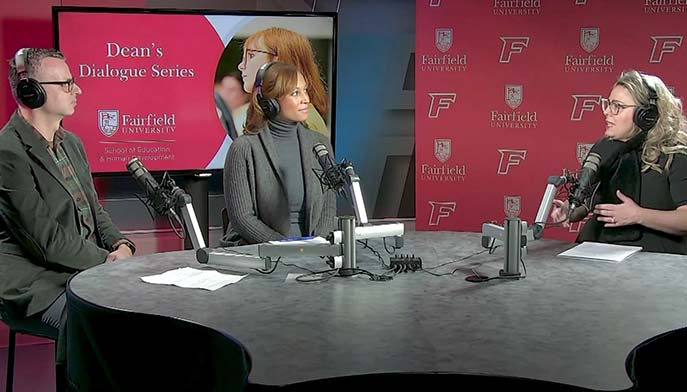 Image of three people sit at a table with microphones, discussing in front of a television screen displaying graphics.